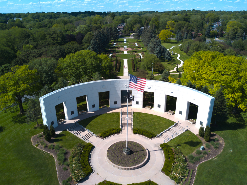 An aerial view of a large park that has a large semicircular memorial for armed forces personnel. You can also see a green lawn, a rose garden, and lots of trees. 