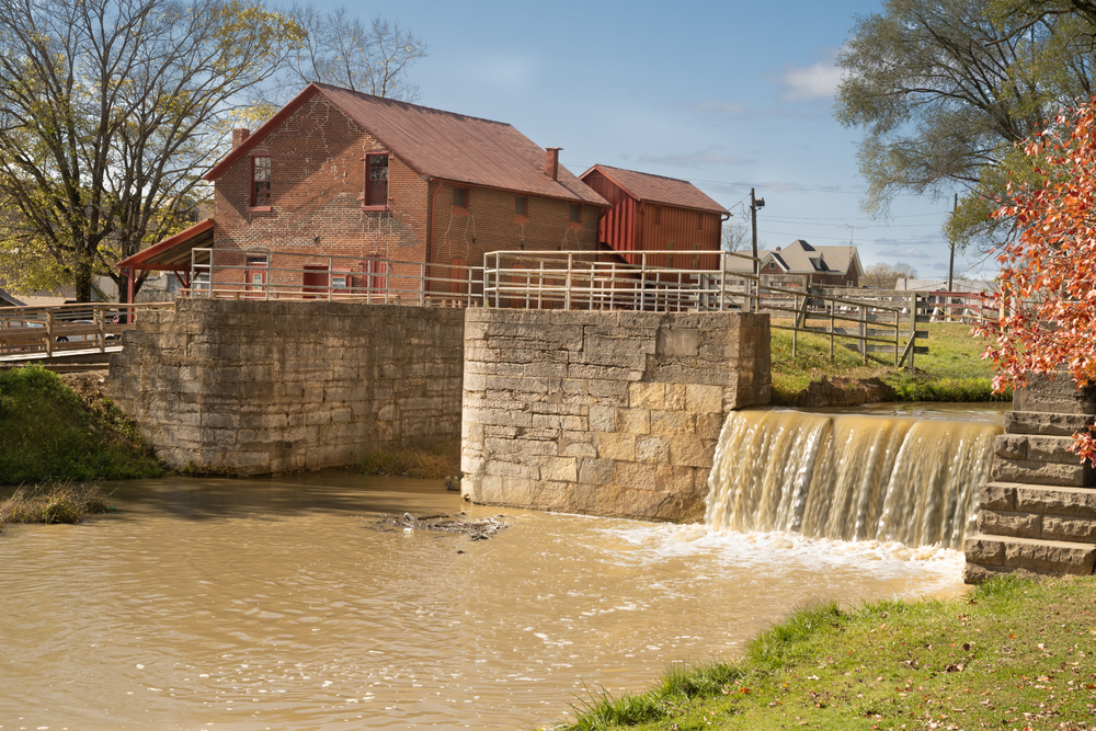 An old grist mill that leads into a river. The mill house is old brick and you can see parts of it that have been patched up. The mill is made out of old stone and creates a small waterfall that runs into a cloudy brown river. 