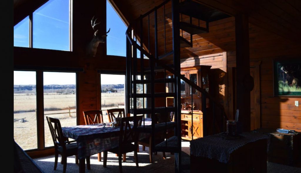 The interior of a log cabins in Nebraska that is partially in shadow. You can see a dining area, a spiral staircase, and windows that look out onto prairies. 