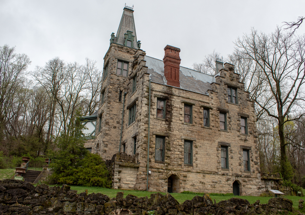 The side of a large stone castle that has Gothic architecture. All around there is a lawn with green grass, dense trees with no leaves, and a stone fence. 