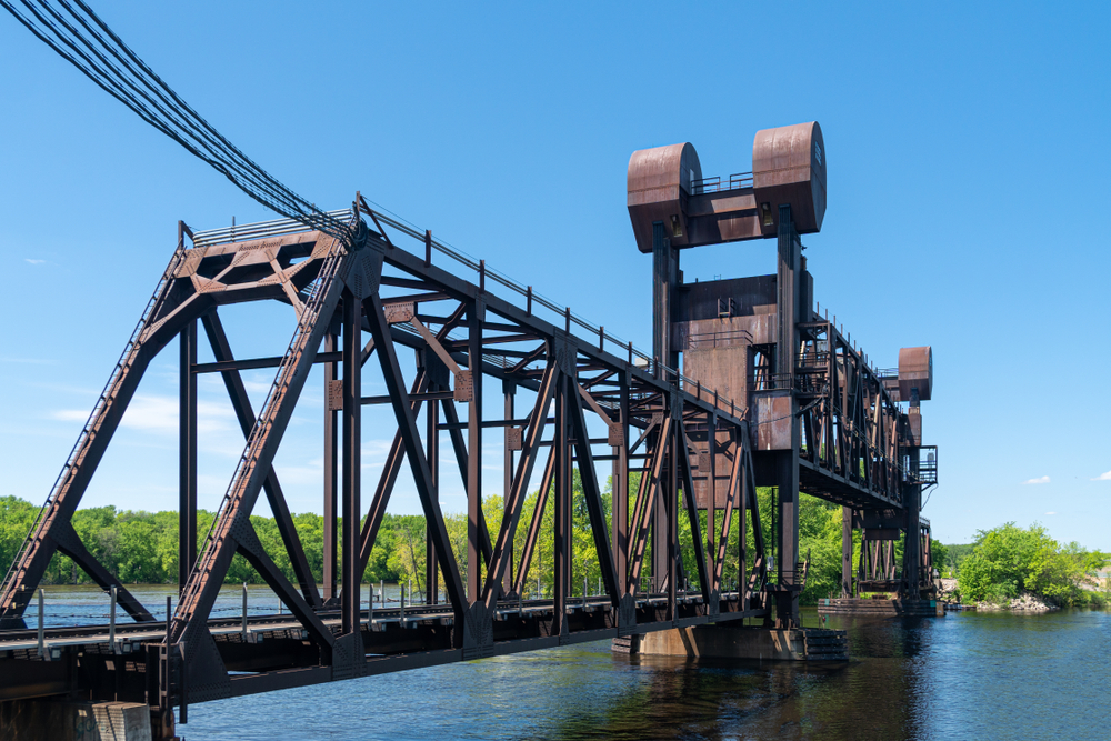 A large metal bridge with lots of connections and wires going over it. It crosses a river where you can see trees on the side of the river bank. 