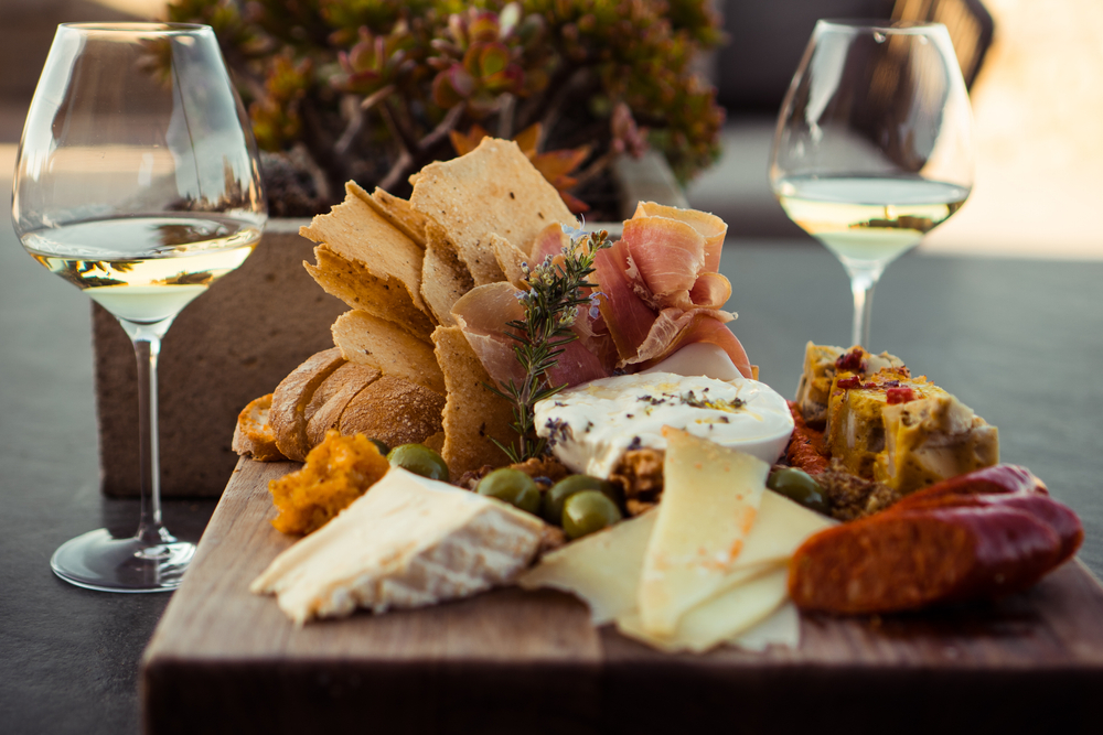 A charcuterie board with chees and ham and two glasses of white wine.