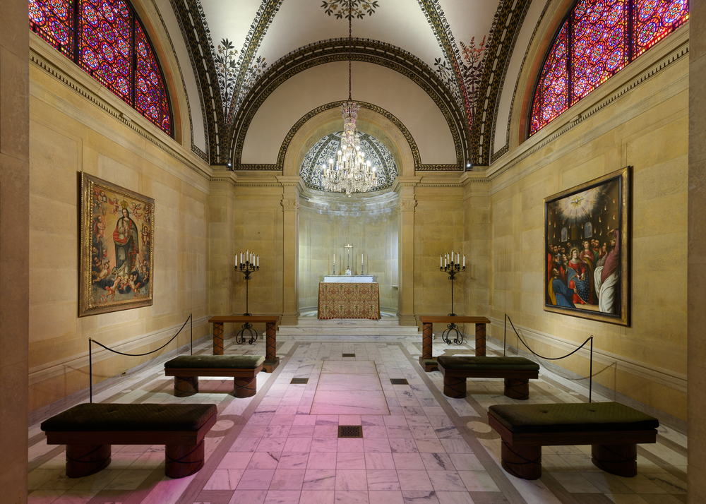 The interior of the chapel of the Saint Cecilia Cathedral in Omaha Nebraska. It has stained glass windows, paintings on the wall, a rounded ceiling, a crystal chandelier, and tile floors with wooden pews.