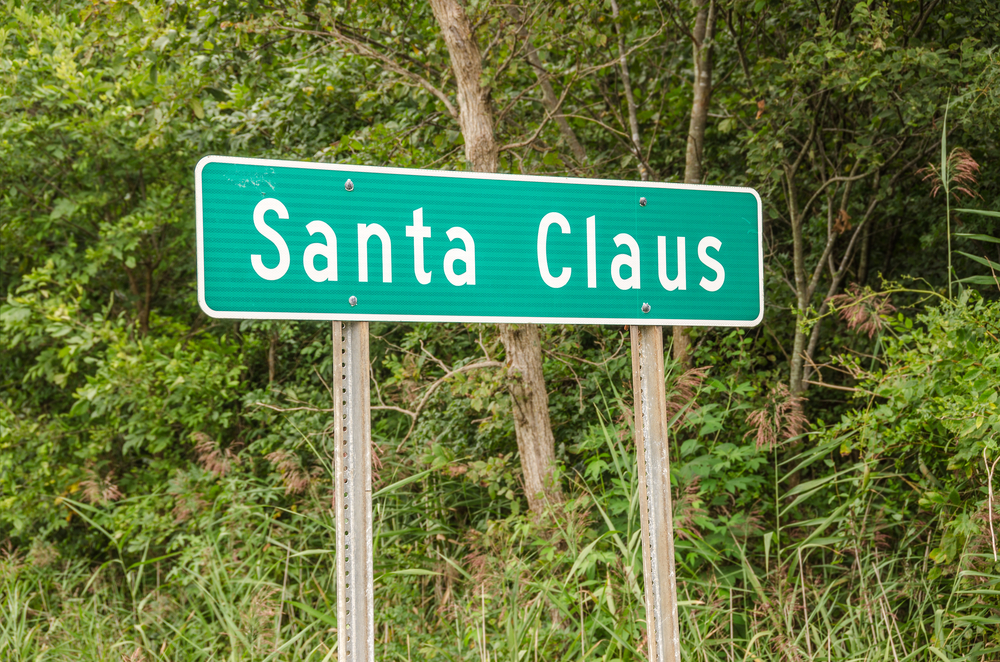 A green sign with 'Santa Claus' on it, welcoming you to the Indiana town of Santa Claus. It is on the side of the road and there are trees behind it. 
