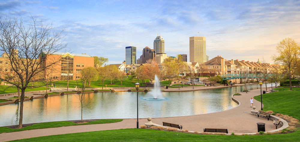 The view of a canal, park, and walkway in Indianapolis, one of the best weekend getaways in Indiana. You can see the city skyline in the distance. 