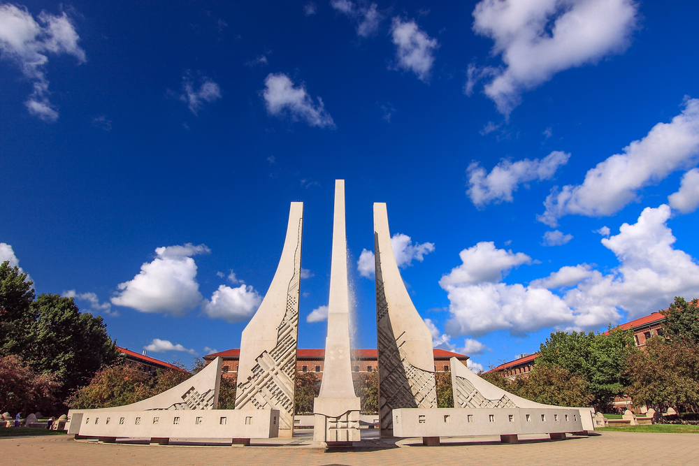 A large sculpture that is separated into three main pieces with two shorter than the one in the middle. Behind it you can see brick buildings with red roofs and a bright blue sky with fluffy clouds. 