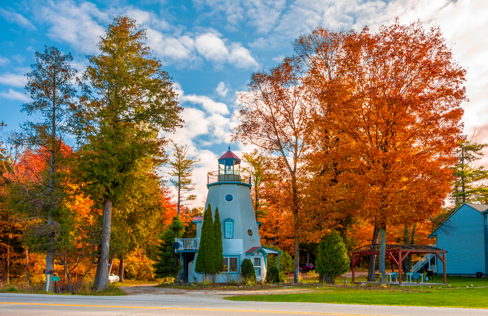 A small lighthouse in a grassy area surrounded by trees with orange, red, yellow, and green leaves. The lighthouse is light gray with a red roof. One of the best Wisconsin road trips stops.