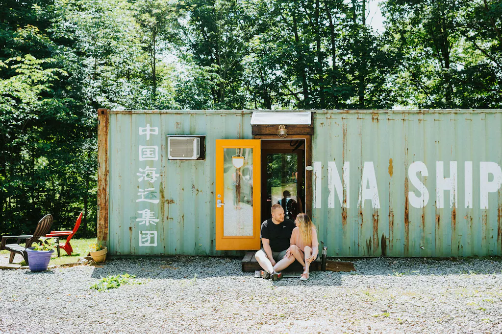 Adult Caucasian couple sitting on doorstep of green rusting container with trees in background and gravel in foreground.