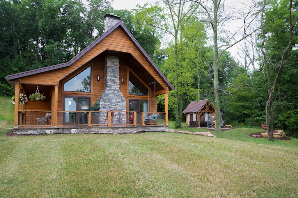 Luxury cabin with stone chimney, front porch, large glass windows. Small covered portico on right.