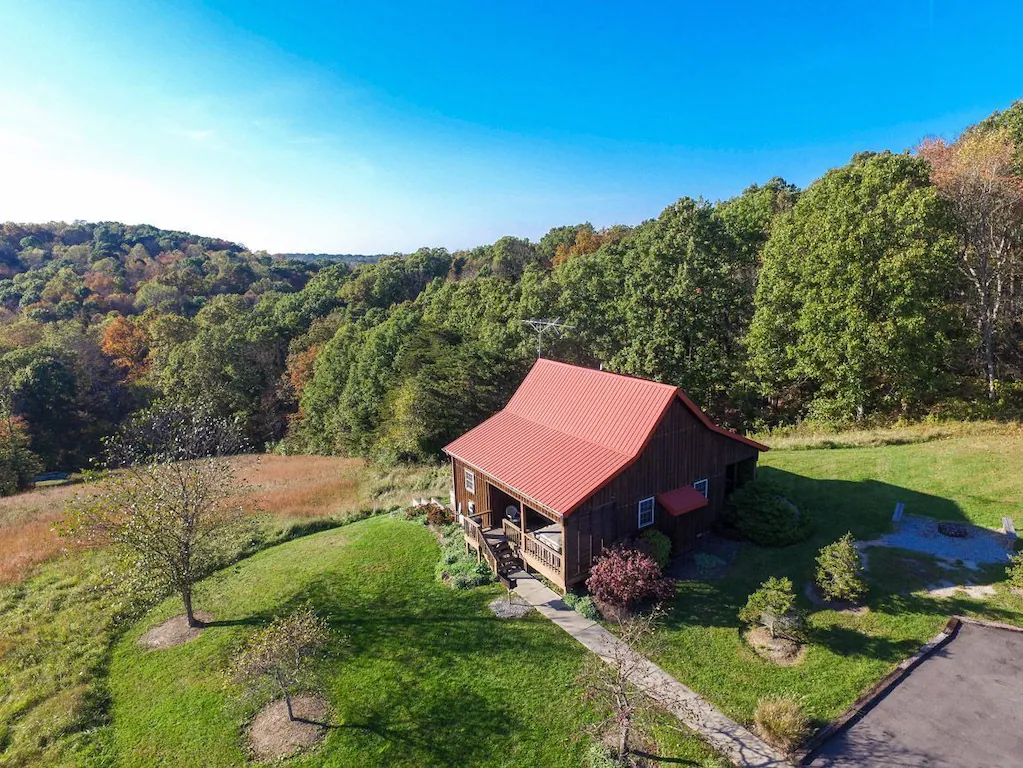 Cabin with red metal roof and front porch site atop hill looking down over forested hillside. VRBO in Ohio.