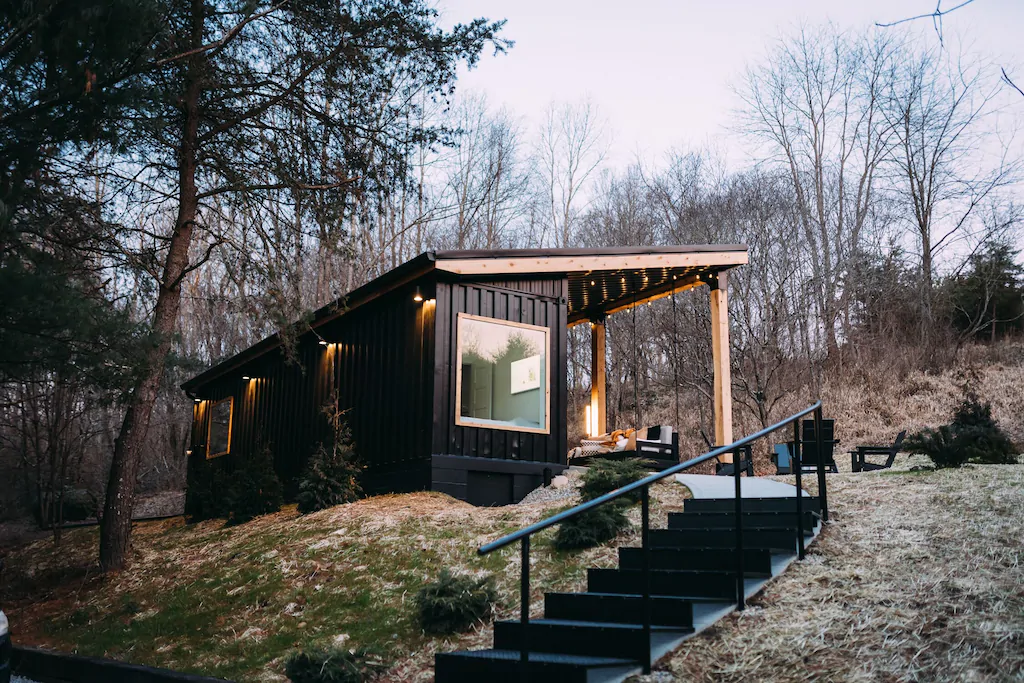 Luxury brown shipping container with large glass window, rooftop covering patio situated on hill. Stairs in foreground.