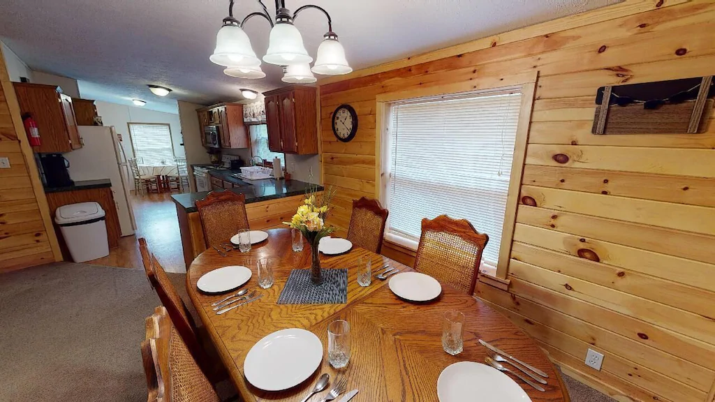 Classic log cabin interior with wood plank walls, dining room table and chairs in foreground, Kitchen in background.
