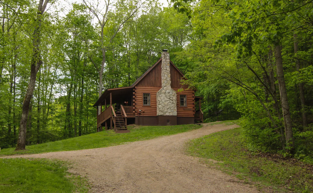 Log cabin sitting on the hill with green forested trees in background and dirt driveway in foreground. 