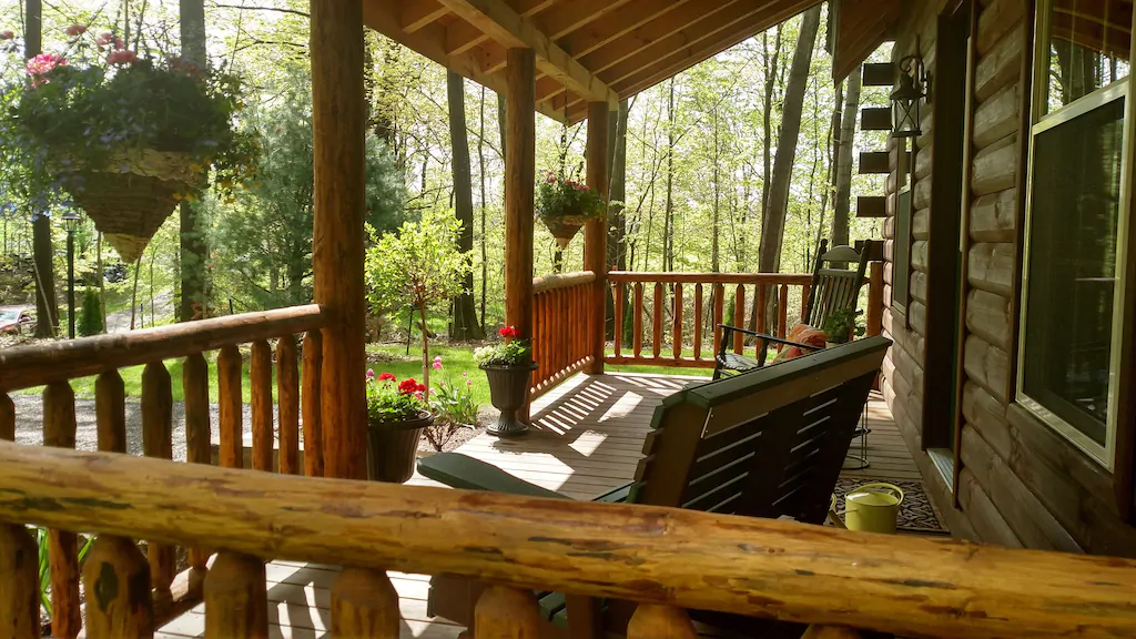 Patio of log cabin looking out to surrounding woods. Wooden chairs on patio with handing baskets with flowers.