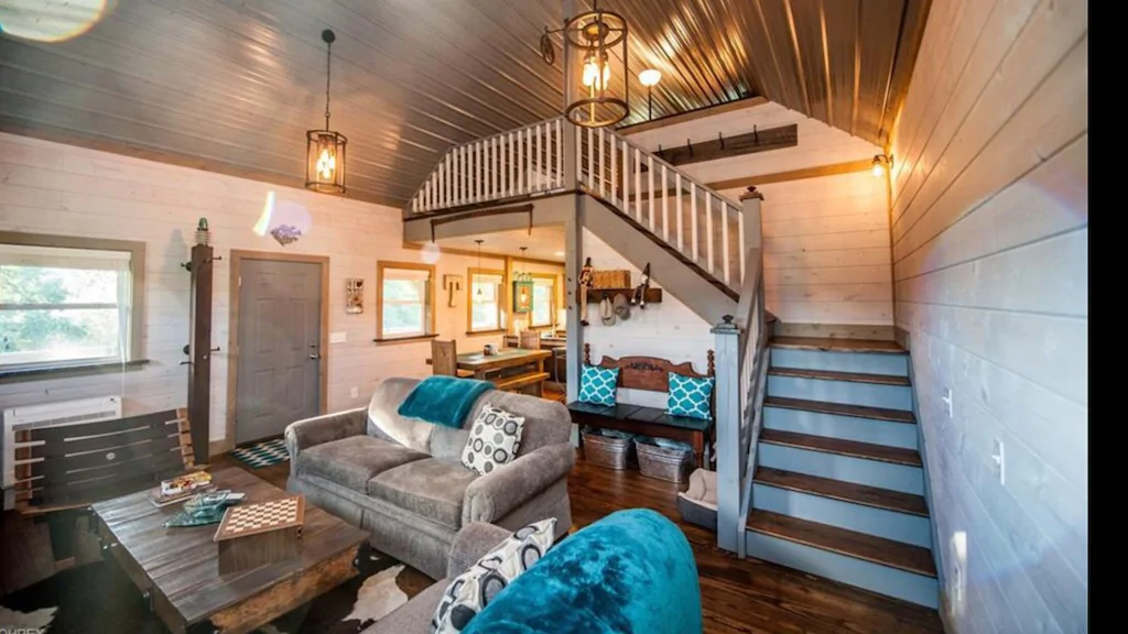 Tastefully furnished open living space with grey couches and turquoise pillows, stairs up to second floor on right and dining table/chairs in background.