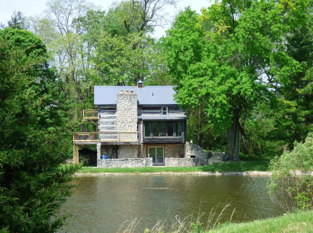 Cabin with stone chimney and wall, with large balcony, surrounded by green trees, lake in foreground.