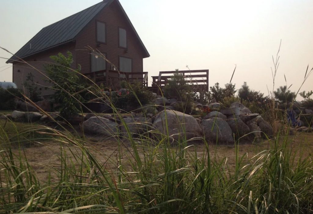 Cabin with metal roof with large balcony with wooden fencing, Large boulders in foreground and tall prairie grass in foreground.