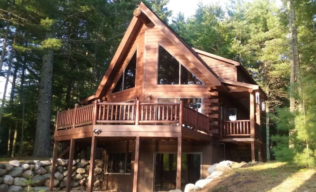 Luxury log cabin with A-frame roof, large wooden balcony, large glass windows, propped up by wooden beams .Green tees in background.
