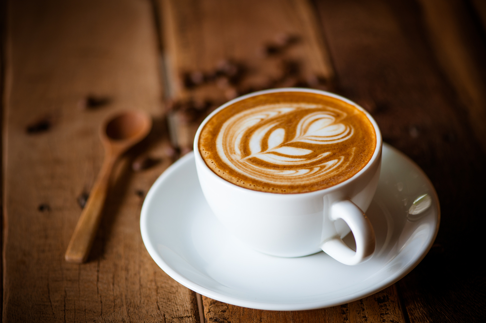 Cup of latte with cream flower decoration, on brown wooden table. Coffee beans and wooden spoon in background. 