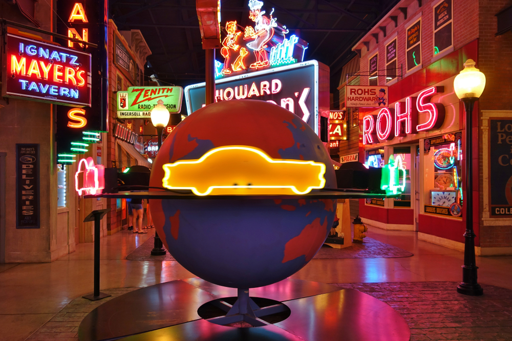 A bunch of neon signs on the side of store front facades in the American Sign Museum. They are all kinds of colors, shapes, and say different things. In the middle is a large globe  with a neon yellow car going around it. 