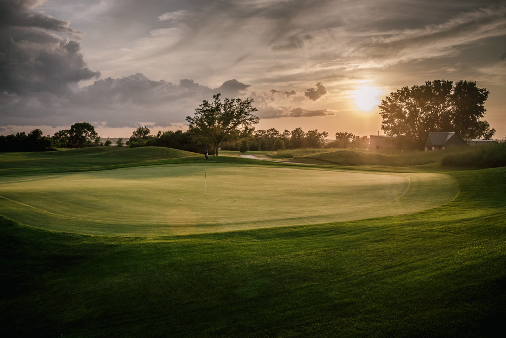 A large golf course at sunrise in Nebraska. There is some fog in the air and clouds are making the sun not so bright. The golf course is very green. 