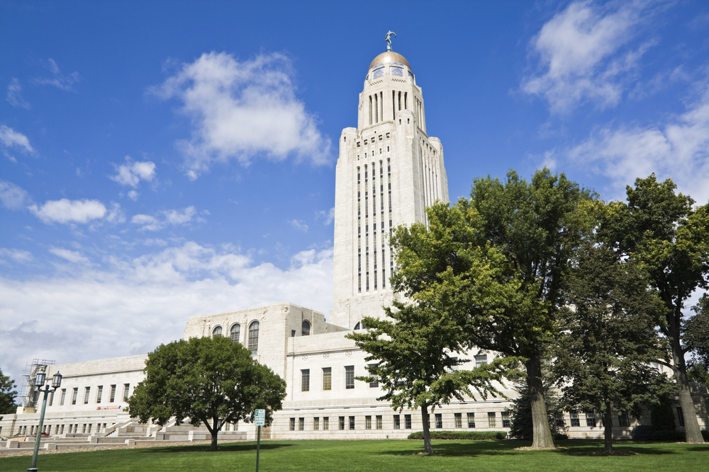 The exterior of the large white Nebraska State Capitol Building. It is long with lots of windows and in the center is a very tall tower with a silver dome and a sculpture on top of it. 