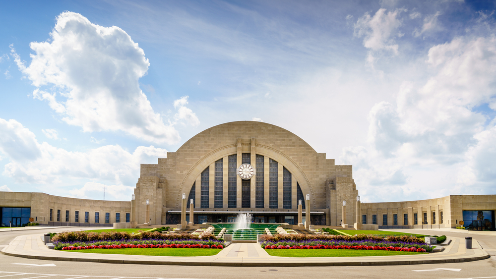 The exterior of the Cincinnati History Museum in the old Union Terminal Station. There are flowers in front of it that are red, white, yellow, and there is a green lawn with a water fountain. 