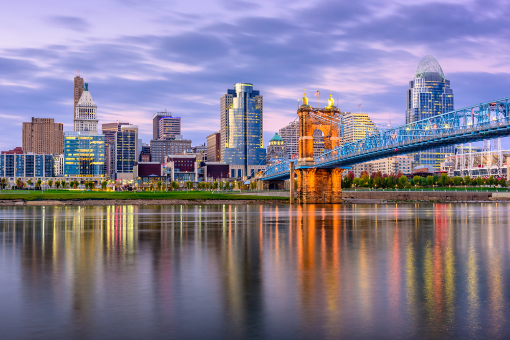 The Cincinnati skyline at twilight across the Ohio River. The buildings are lit up, the sky is purple and pink, and you can see the lights of the buildings in the river.