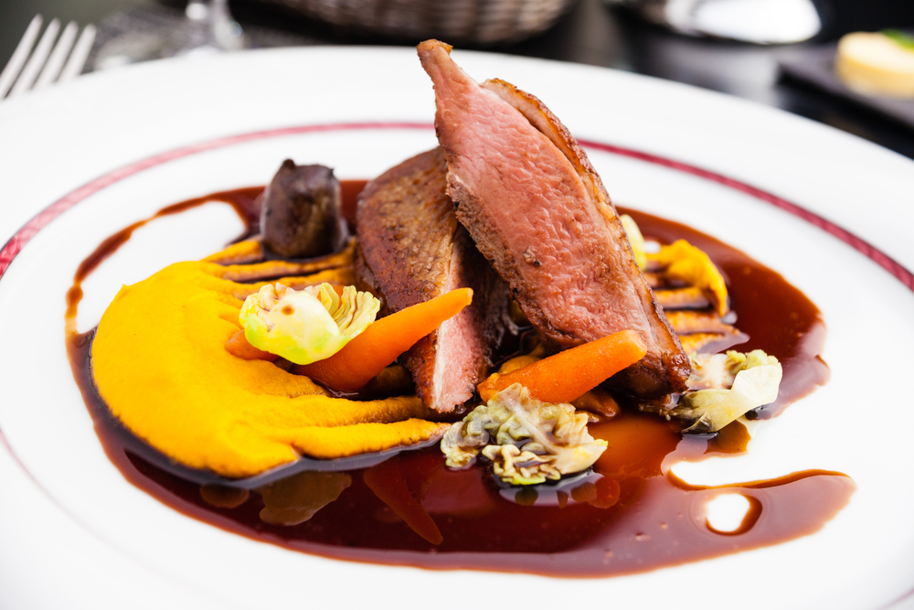 An ornately plated French dish. The plate is white and there is some sort of pureed squash, grilled duck, some other vegetables, and a brown sauce. 