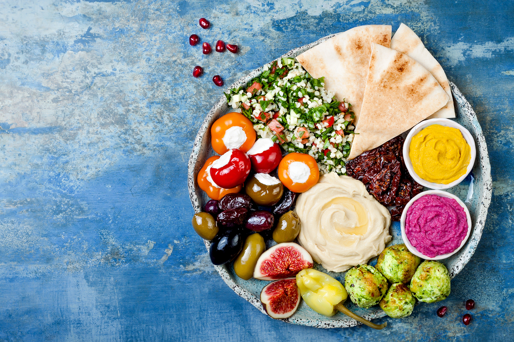 Looking down at a large platter of Greek food on a blue white-washed table top. The platter has hummus, roasted brussels sprouts, stuffed tomatoes, olives, pita bread, and a couscous salad. Its similar to what you can find at one of the best Greek restaurants in Lincoln Nebraska. 