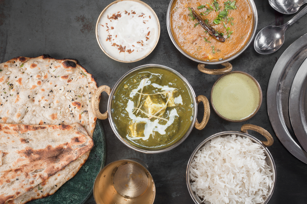 Looking down at a black table covered in dishes full of traditional Indian cuisine. There is paneer in a green sauce, an orange curry, a white yogurt sauce, a green yogurt sauce, biryani rice, and naan bread. 