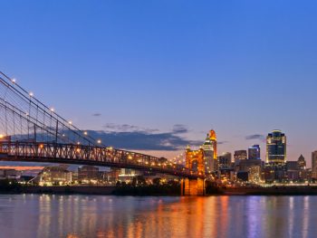 A view of the John A. Roebling Suspension Bridge, one of the coolest things to do in Cincinnati. It is twilight and the bridge is lit up. You can also see a city lit up on the other side of the bridge.