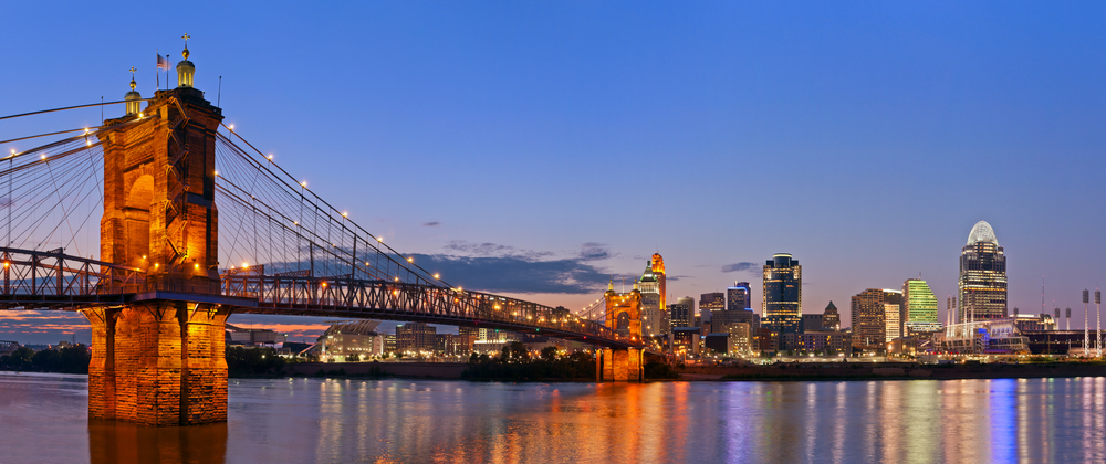 A view of the John A. Roebling Suspension Bridge, one of the coolest things to do in Cincinnati. It is twilight and the bridge is lit up. You can also see a city lit up on the other side of the bridge.