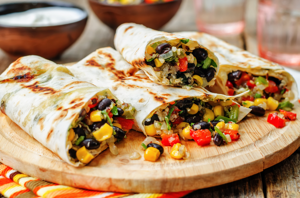 A wooden platter with grilled burritos overflowing with a variety of vegetables. You can see corn, black beans, chopped tomato, chopped pepper, herbs, and quinoa. 