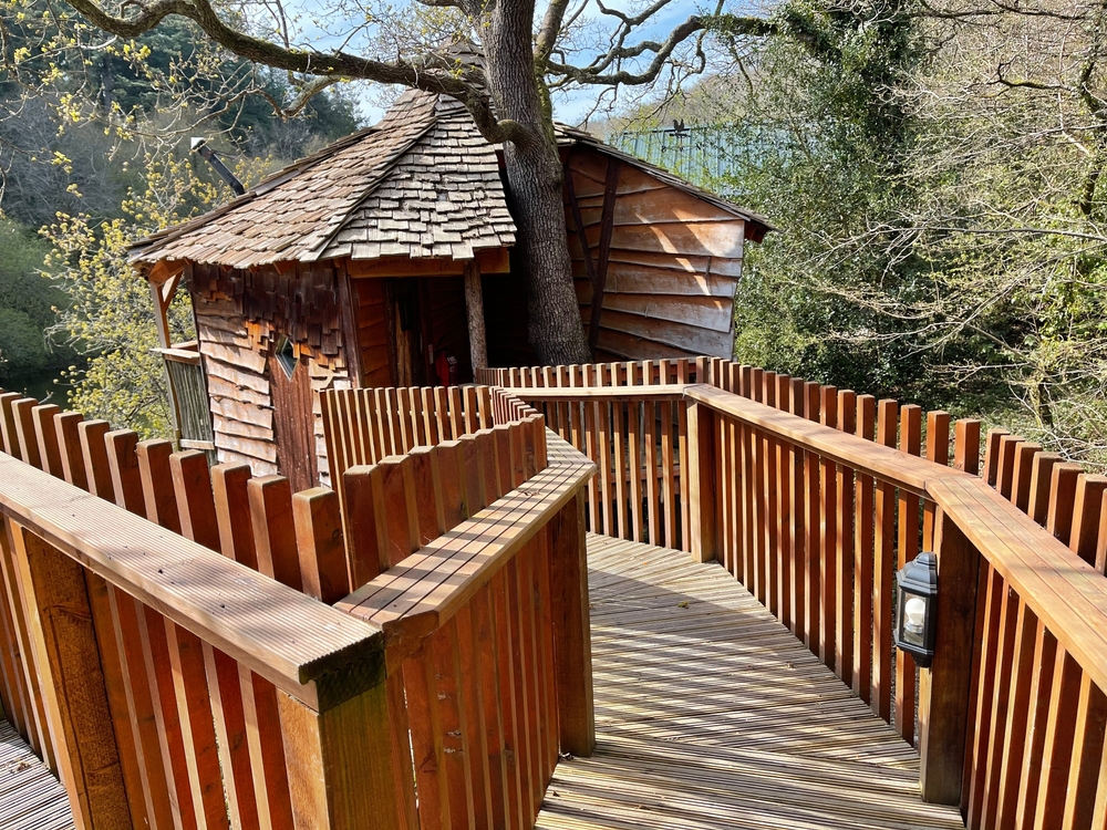 A treehouse like the one in the Mt. Airy Forest, one of the best things to do in Cincinnati Ohio. There is a wooden walkway, a small treehouse with wooden shingles, and it is surrounded by trees. 