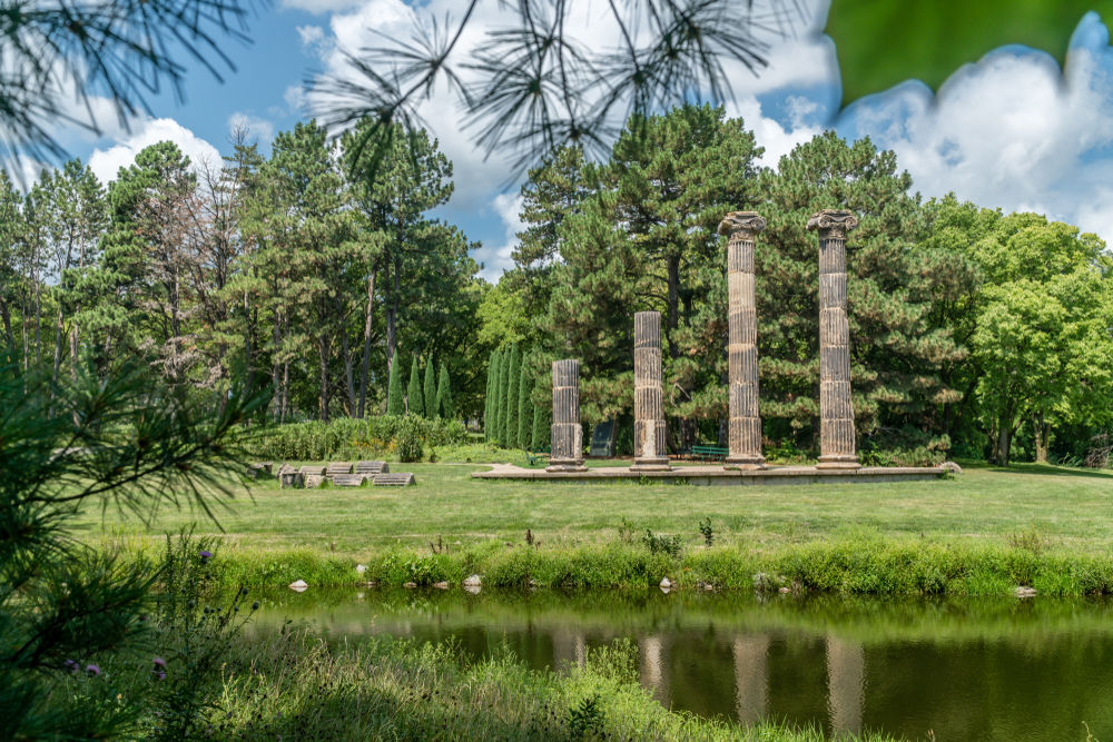 Looking from the bank of a pond across the pond at a park. There are old columns on a concrete slab, trees, a lawn, and shrubs. It's one of the best things to do in Lincoln NE.