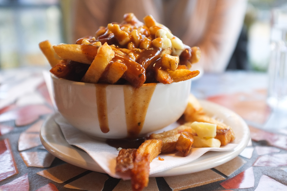 A white bowl on a saucer on a mosaic table. The bowl is overflowing with French fries covered in poutine. 