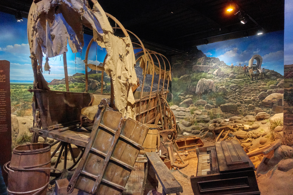 A display of a pioneer wagon that is torn apart in a museum in Lincoln NE. Behind it there is a painting of pioneers making their way west. 