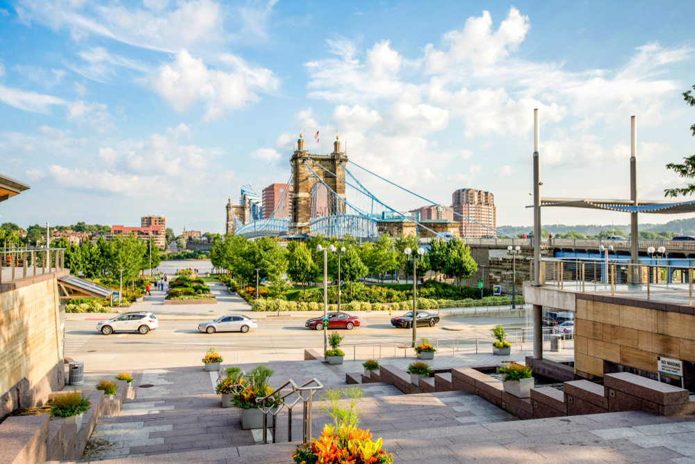 The view of the Smale Riverfront Park near the John A. Roebling Suspension Bridge in Cincinnati. There are trees, shrubs, stone walkways, and stone steps. 