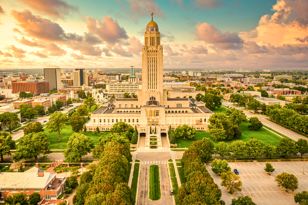 The Lincoln Nebraska skyline on a sunny day. You can see the large capitol building, the green park around it, lots of trees, buildings, and roads. The sky is full of fluffy clouds. 