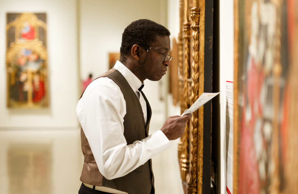 A man in a white shirt and brown vest looking closely at art in ornate gold frames in an art museum. 