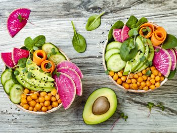 Two bowls on a light gray wood table. The bowls are vegan bowls from one of the vegan restaurants in Omaha. You can see sliced avocado, cucumbers, squash, spinach, chickpeas, and a pink fruit in the bowl.