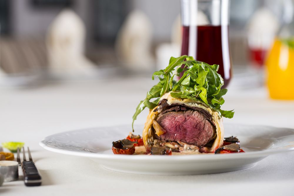 A white plate on a fancy table with a white tablecloth. On the plate there is a slice of beef wellington with greens on top of it. You can also see a creamy sauce and roasted tomatoes. On the table you can also see a glass of red wine.