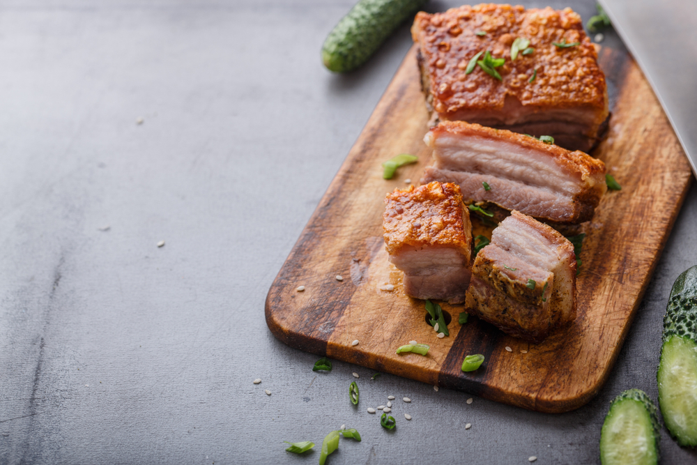 Crispy pork belly on a wooden chopping block. It's similar to a dish you can find at one of the best restaurants in Detroit. You can also see pickles and a knife near the cutting board.