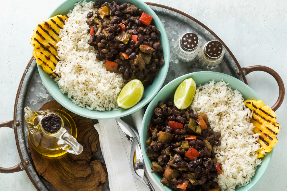 An aluminum tray with two bowls of food. The bowls are full of black beans, rice, grilled pineapple, and a lime wedge. It's similar to a meal you can find in restaurants in Detroit.