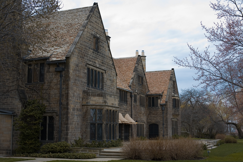 The exterior of the ornate Ford House. It is made of different colored brick and is very large. Around it you can see trees and bushes with no leaves. 