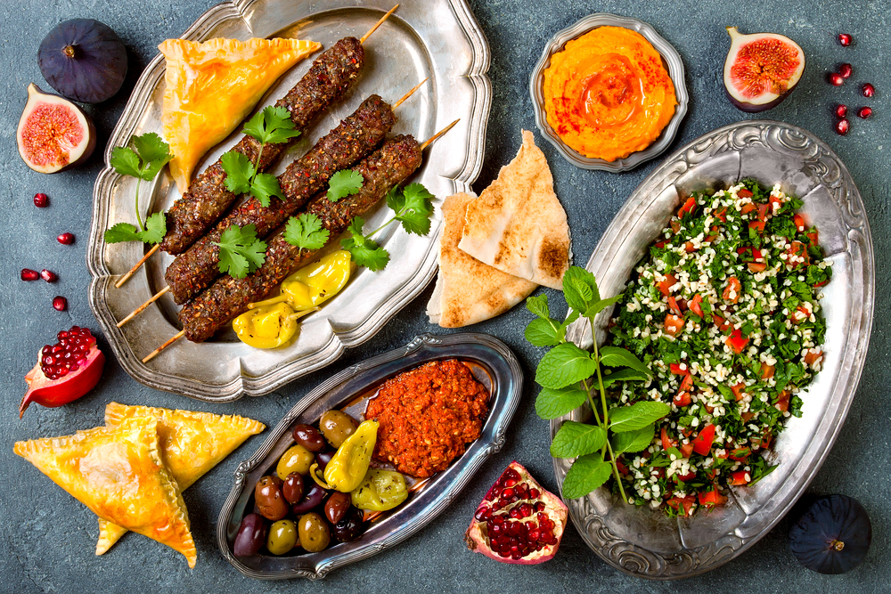 A spread of Middle Eastern food. There is kabobs, a quinoa salad, hummus, pita, pomegranates, and figs. The foods are on silver dishes. Its similar to meals you find in restaurants in Detroit.