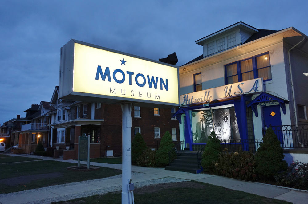 The exterior of the Motown Museum. There is a large sign on the yard of the old white house that is lit up that says 'Motown Museum'. The house is white with cobalt blue trim. 