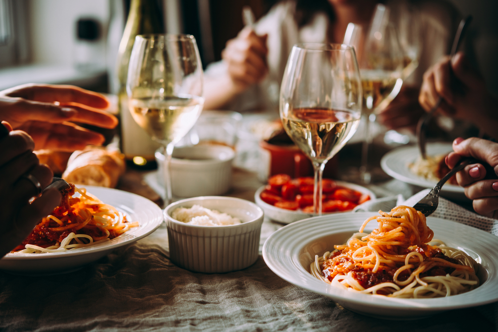 Three people eating pasta around a table with wine