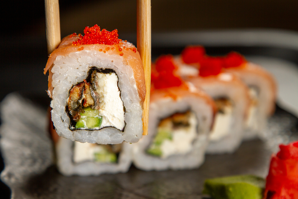 Sushi in chopsticks with more sushi on a plate behind